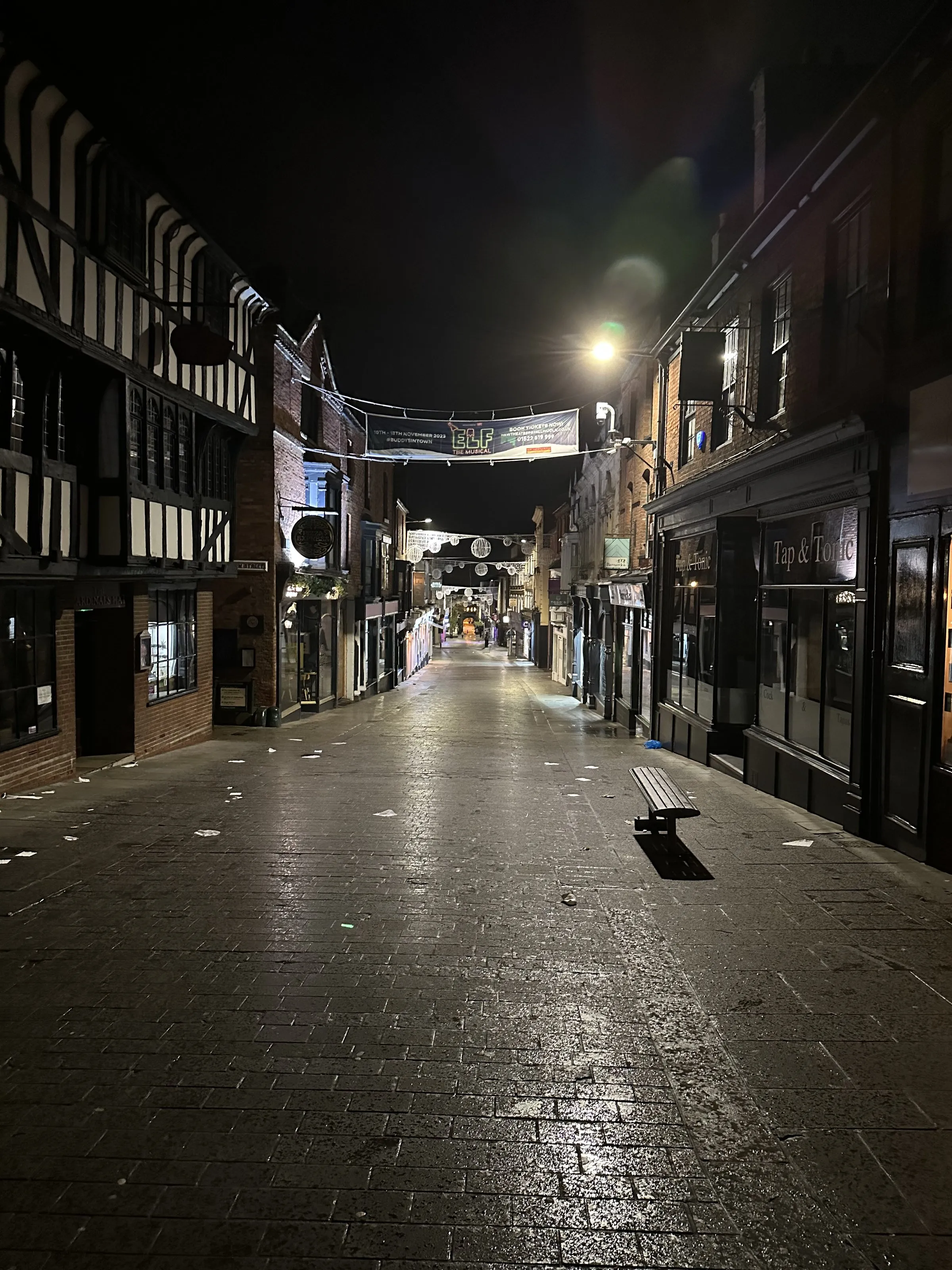 An empty cobbled street of timber-framed buildings under street lamps before dawn