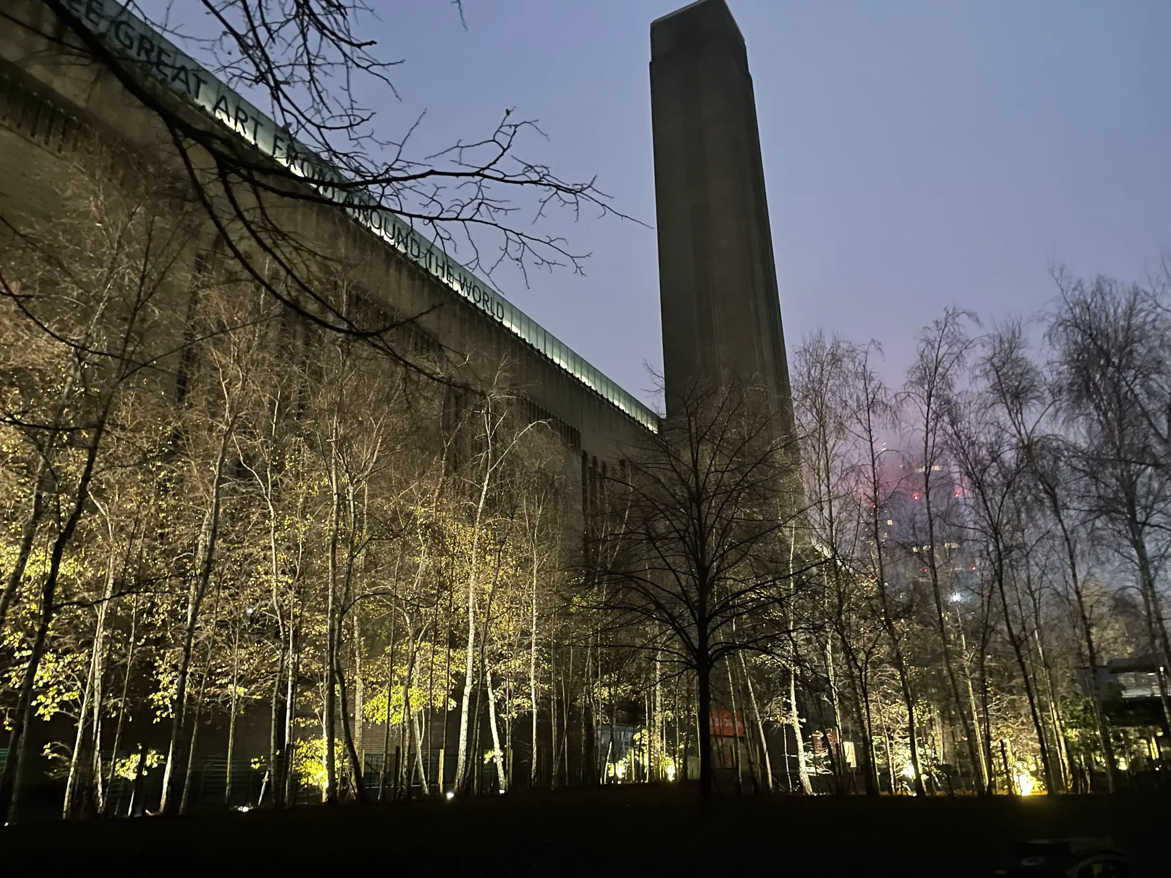 Tate Modern seen from below before dawn, its chimney rising above illuminated birch trees