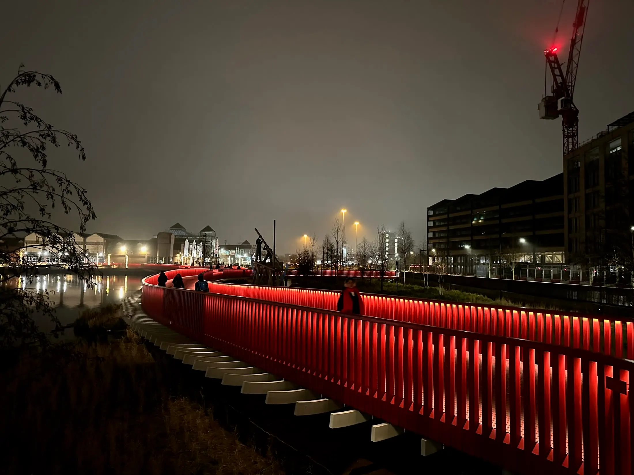 The red footbridge at Canada Water at night, its rail lit from within, people crossing towards the lights