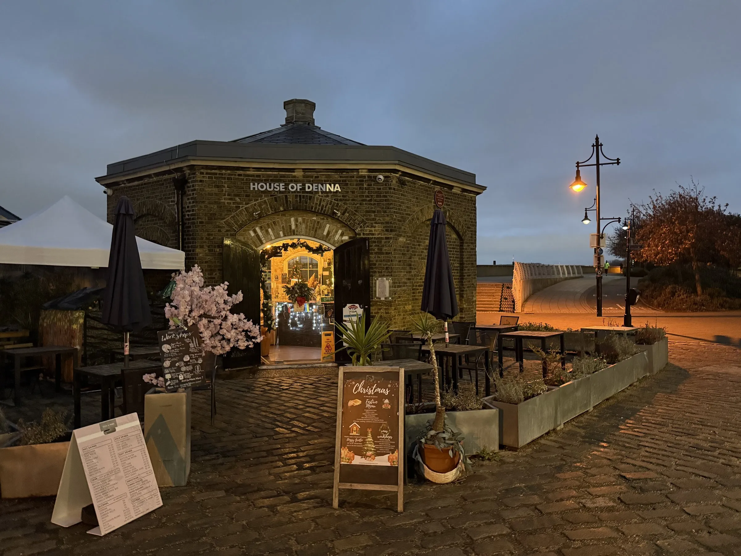 An old brick guardhouse at the Royal Arsenal, now a café, its doors open on fairy lights across empty cobbles before sunrise